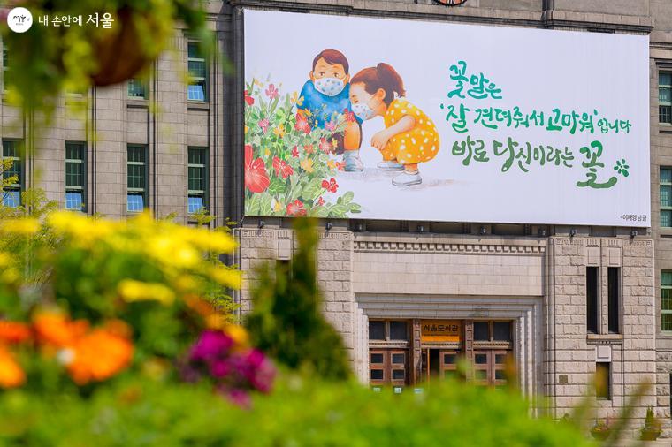 서울광장 잔디마당에 피어있는 꽃들 사이로 바라본 서울도서관 꿈새김판. 다정한 인사를 건네는듯하다 ⓒ문청야