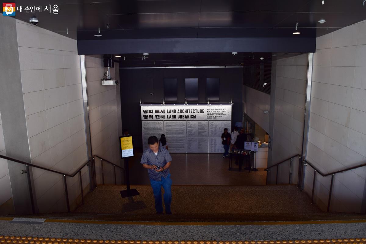 서울도시건축전시관 입구. 입구에는 지하로 내려가는 계단이 보이며 전시를 소개하는 현수막이 크게 부착되어 있다. ©이명은