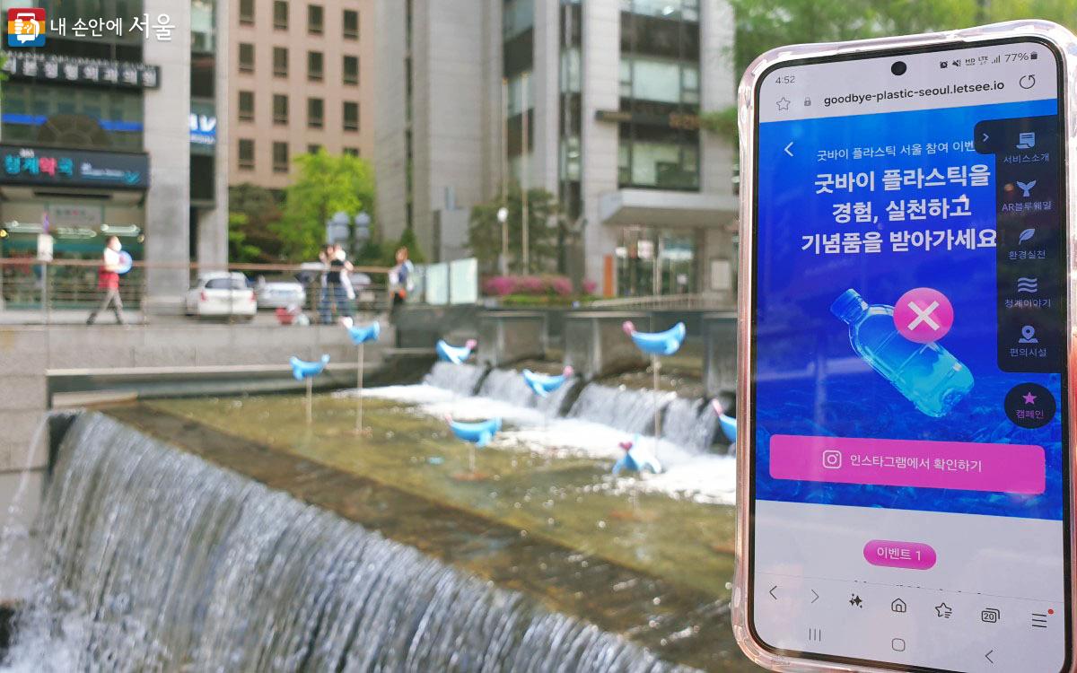 푸른 고래 조형물 오른쪽에서 스마트폰으로 굿바이 플라스틱 서울의 이벤트 안내 창을 보고 있다. ©송지혜
