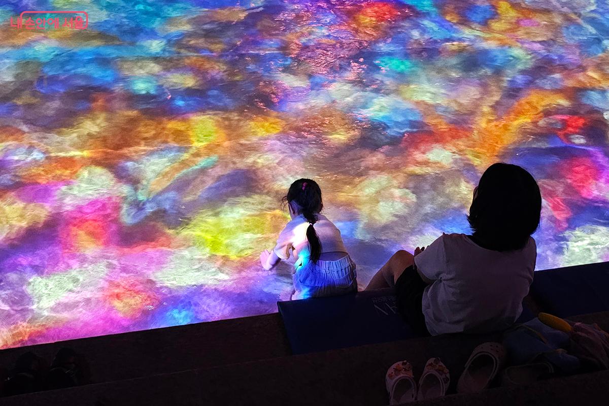 알록달록한 파스텔 톤의 영상이 비추는 청계천 물결 위로 어린아이가 손을 뻗어 물장구를 치고 있다. ©정향선