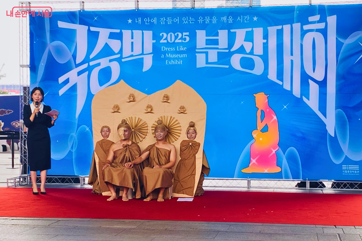 '소분모임' 팀이 무대 위에서 '이불병좌상'을 표현하고 있다. 4명의 참가자가 갈색 천을 두르고 부처의 모습을 한 채 앉아 있고, 그 뒤에는 커다란 광배 모양의 배경이 있다. 여성 사회자가 옆에서 마이크를 들고 소개하고 있다. ©양정화