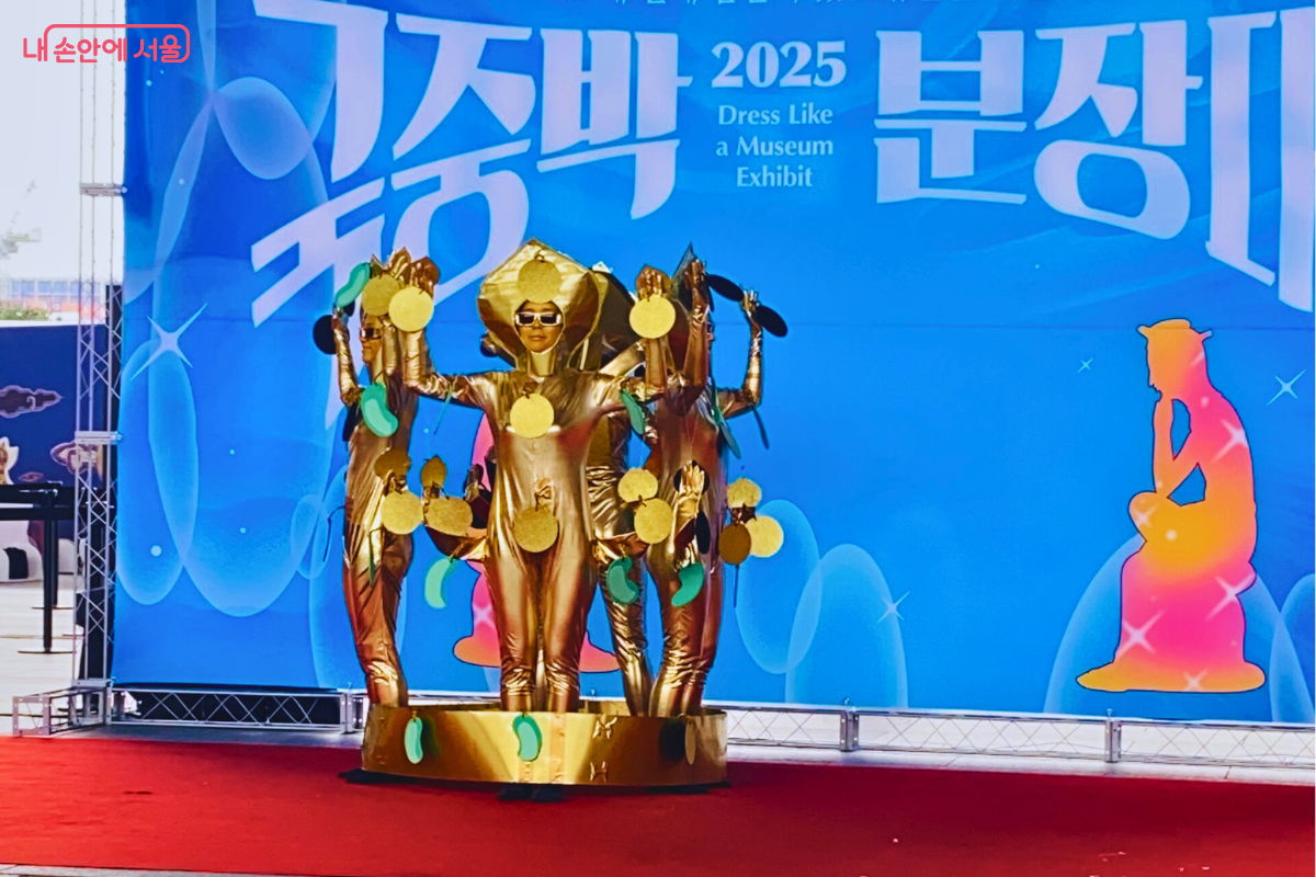 4명의 참가자가 금색 의상을 입고 서봉총 금관을 표현하는 퍼포먼스를 하고 있다. 가운데 참가자는 선글라스를 끼고 있으며, 네 사람 모두 금관의 장식을 표현한 소품을 들고 위풍당당한 포즈를 취하고 있다. ©양정화