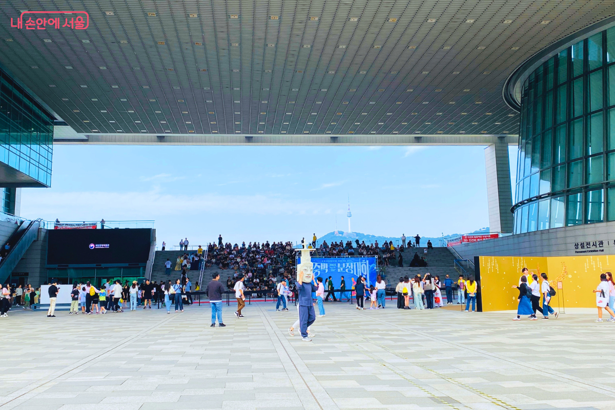 국립중앙박물관 열린마당의 넓은 광장 모습. 계단에는 행사를 기다리는 많은 시민이 앉아 있고, 광장 곳곳에서도 시민들이 삼삼오오 모여 사전 행사를 즐기고 있다. 멀리 남산타워가 보인다. ©양정화