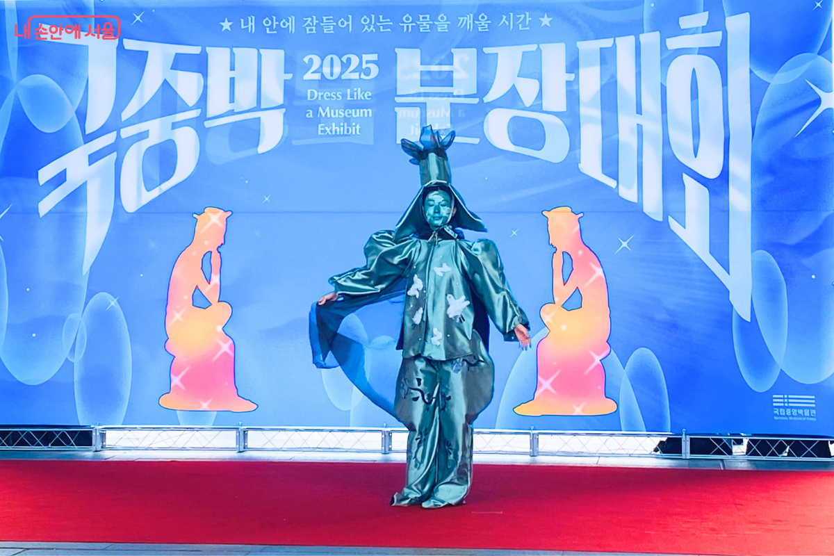 '국중박 분장대회' 무대 위에서 참가자 장 씨가 고려청자를 표현하고 있다. 청록색의 광택이 나는 의상을 입고 있으며, 머리 위에는 병의 주둥이 모양 장식이 있다. 옷에는 청자 상감 무늬 모란국화문과 무름 무늬가 그려져 있고, 팔을 벌린 채 퍼포먼스를 하고 있다. ©양정화