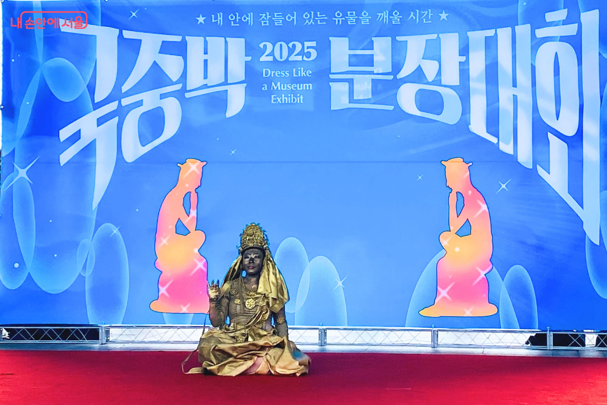 '국중박 분장대회' 무대 위에서 참가자 장 씨가 고려청자를 표현하고 있다. 청록색의 광택이 나는 의상을 입고 있으며, 머리 위에는 병의 주둥이 모양 장식이 있다. 옷에는 청자 상감 무늬 모란국화문과 무름 무늬가 그려져 있고, 팔을 벌린 채 퍼포먼스를 하고 있다. ©양정화