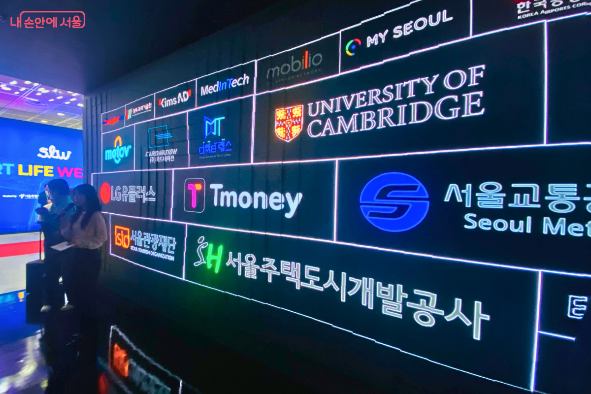 스마트라이프위크에 참여한 여러 기업과 기관의 로고가 표시된 대형 디지털 월. 케임브리지 대학교, 서울교통공사, 티머니 등 국내외 유수 기관들의 로고가 나열되어 행사의 높은 위상과 폭넓은 협력 관계를 보여준다.