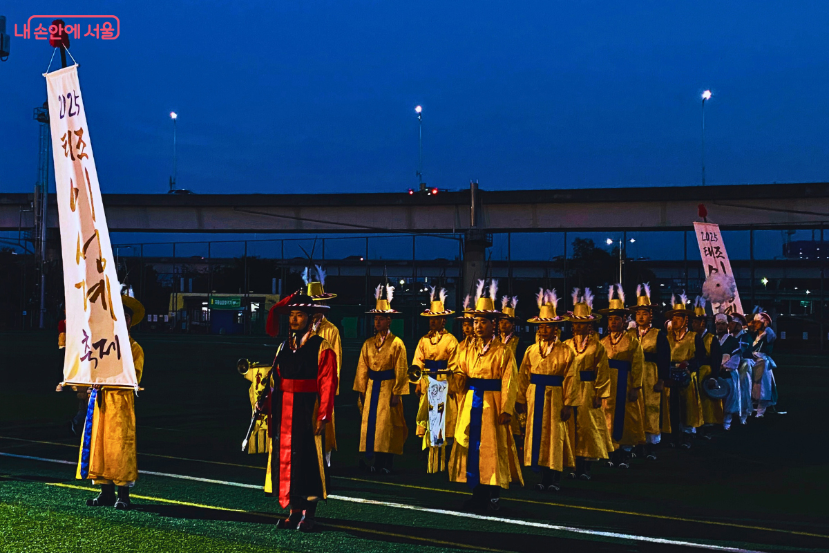 해가 진 저녁, 살곶이 체육공원 잔디밭에서 '태조 이성계 축제' 깃발을 든 공연단을 선두로, 노란색 전통 의상을 입은 평화취타대와 연희앙상블 ‘비단’ 단원들이 '판굿' 공연 입장을 위해 정렬해 서 있다.