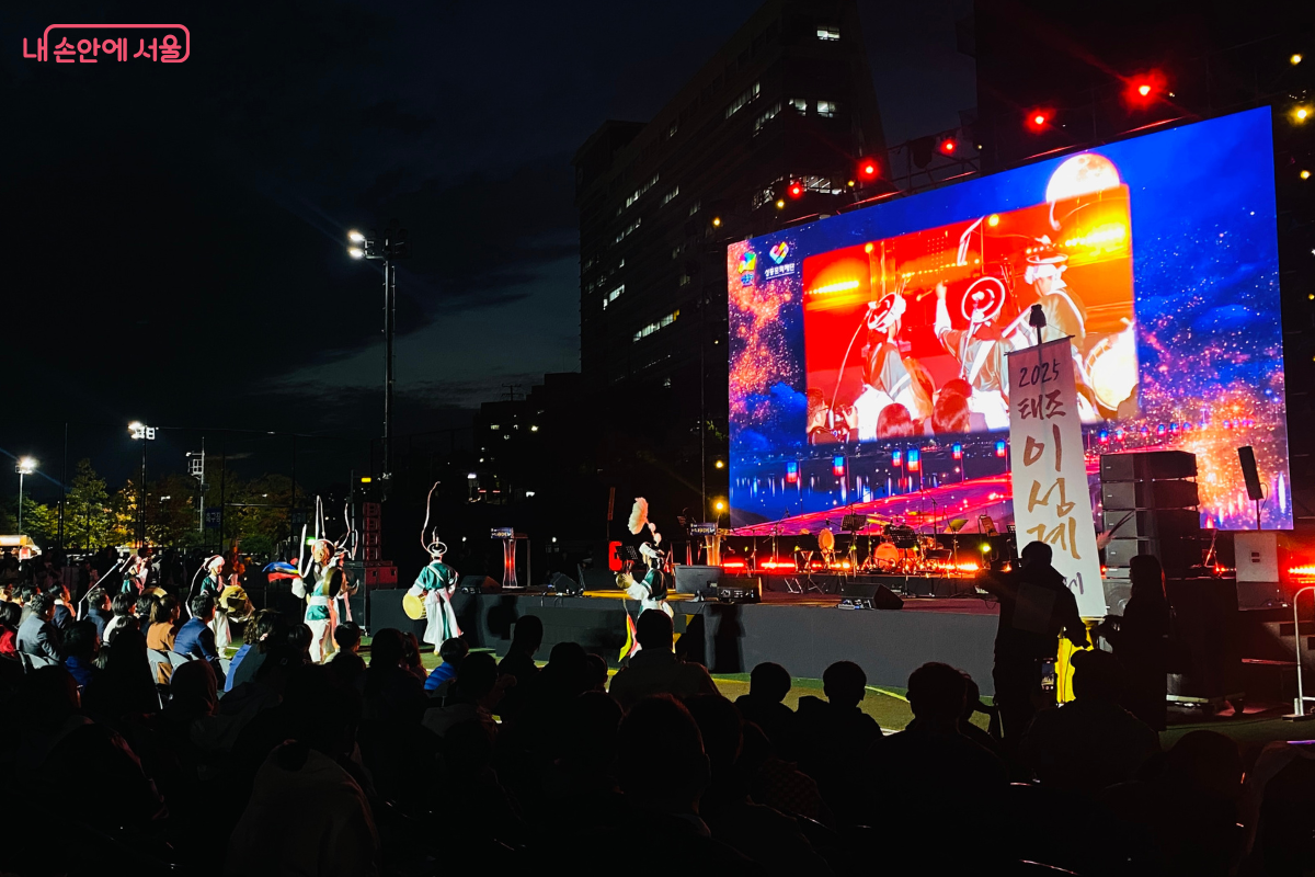 연희앙상블 ‘비단'이 메인 무대와 잔디밭에서 판굿 공연을 펼치고 있다. 대형 스크린에는 공연 모습이 실시간으로 중계되고 있으며, 많은 관객이 공연을 지켜보고 있다.