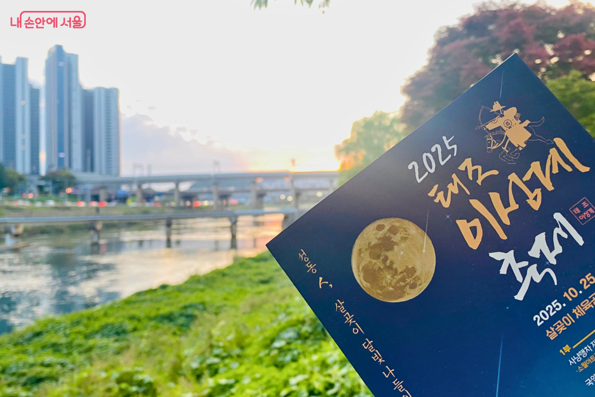  '2025 태조 이성계 축제' 안내 책자를 손에 들고 중랑천 너머로 보이는 살곶이 다리를 배경으로 촬영한 사진. 해 질 녘 노을과 함께 태조와 태종의 이야기가 깃든 살곶이 다리(보물 제1738호)의 모습이 보인다.