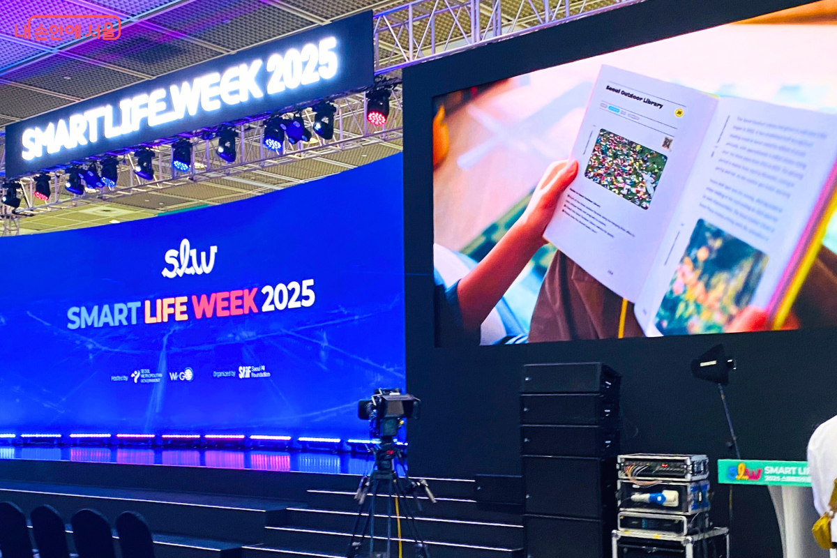 'SMART LIFE WEEK 2025' 행사장 무대와 대형 스크린의 모습. 무대 위 스크린에는 행사 로고가 선명하게 표시되어 있고, 옆 스크린에는 한 사람이 책을 읽는 장면이 나오고 있다. 행사의 시작을 알리는 공식적인 분위기를 연출한다.