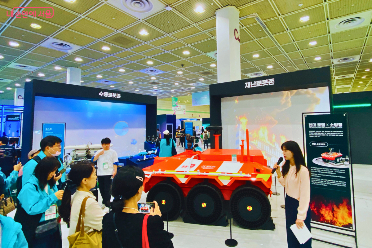 현대 로템과 소방청이 협력하여 개발한 재난 대응 로봇이 전시된 부스. 주황색의 견고한 다목적 로봇 주위로 시민들이 모여 도슨트의 설명을 듣고 있다. 화재 진압 등 인간이 접근하기 어려운 재난 현장에서 활약할 로봇 기술에 대한 높은 관심을 보여준다.