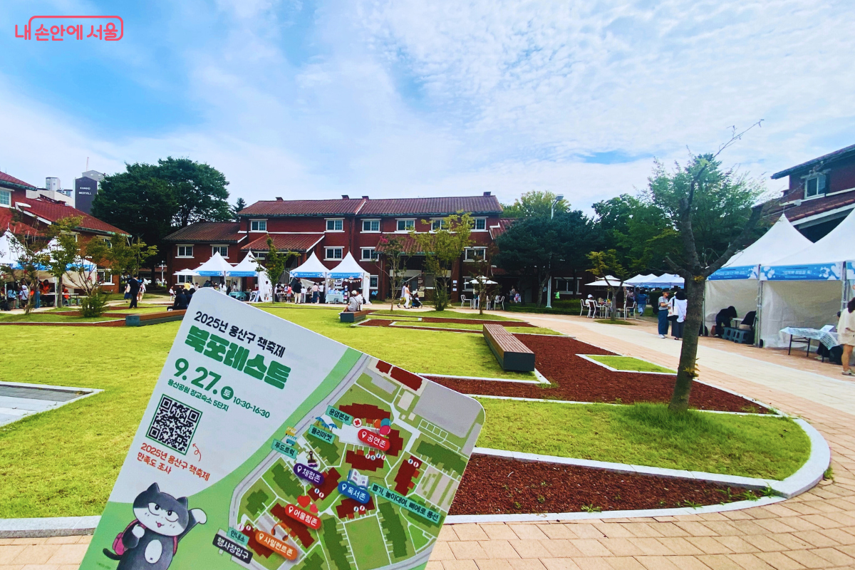 한손에 '북포레스트' 축제 안내도를 들고 행사장 전경을 바라보는 구도의 사진. 푸른 잔디와 붉은 벽돌 건물을 배경으로 여러 체험 부스 텐트와 시민들의 모습이 보인다. ©양정화