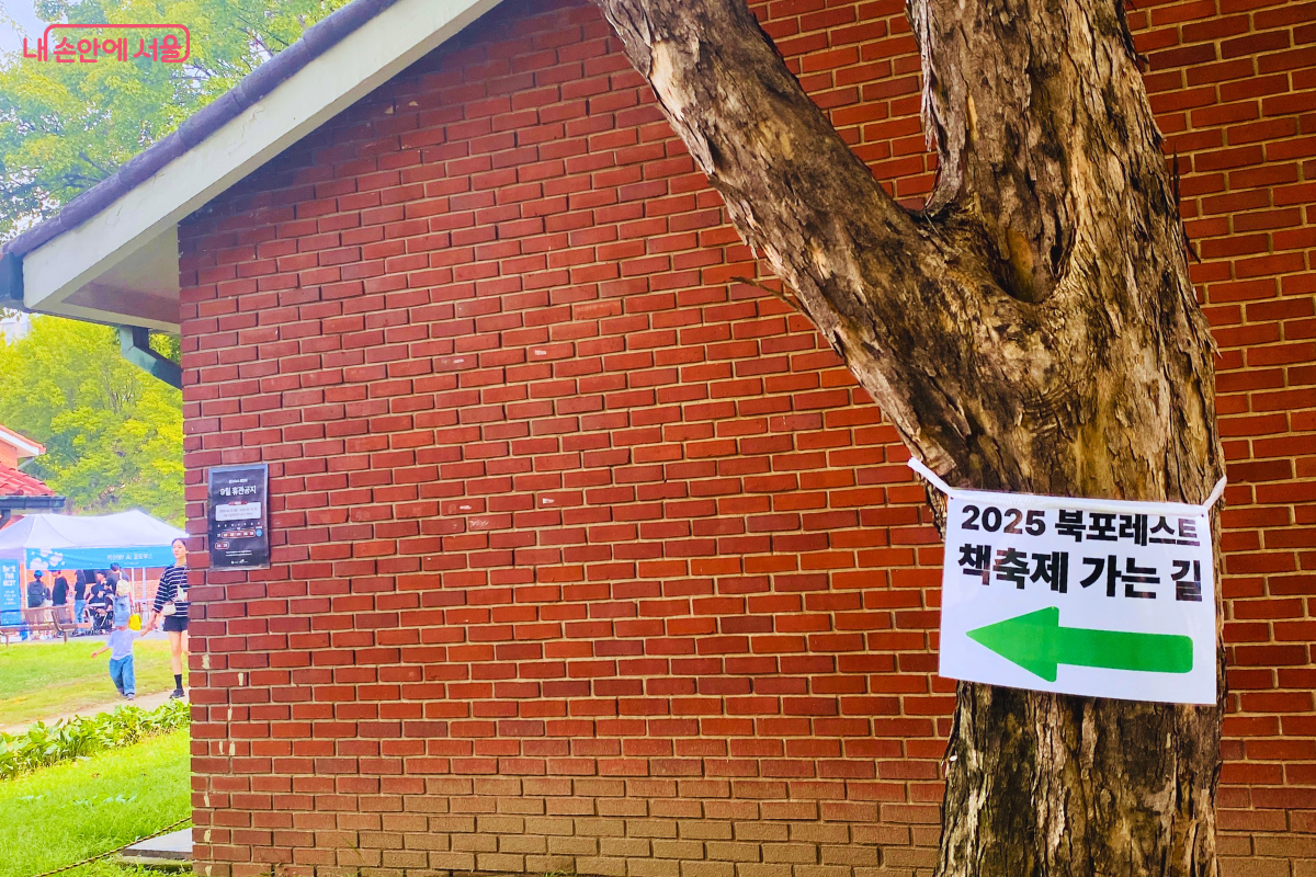 붉은 벽돌 건물 옆 나무 기둥에 '2025 북포레스트 책축제 가는 길'이라고 적힌 안내문이 붙어 있다. 초록색 화살표가 행사장 방향을 가리키고 있다. ©양정화