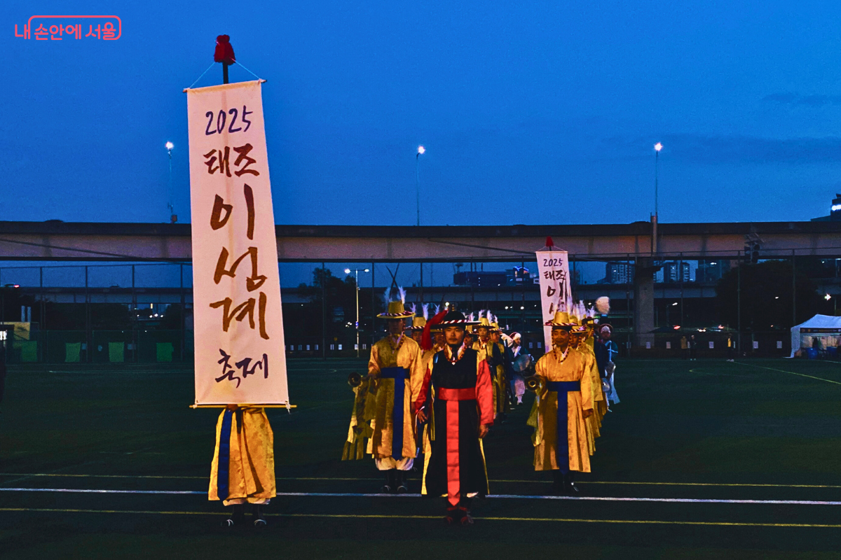 어두워진 축구장에서 '2025 태조 이성계 축제'라고 쓰인 깃발을 든 기수 두 명과 전통 복장을 한 공연단이 입장을 준비하며 정면을 바라보고 있다.