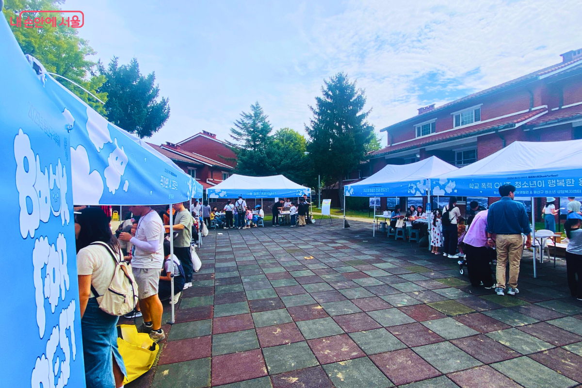 광장에 여러 개의 체험 부스가 설치되어 있다. 많은 시민이 각 부스를 둘러보거나 체험에 참여하며 활기찬 축제 분위기를 만들고 있다.