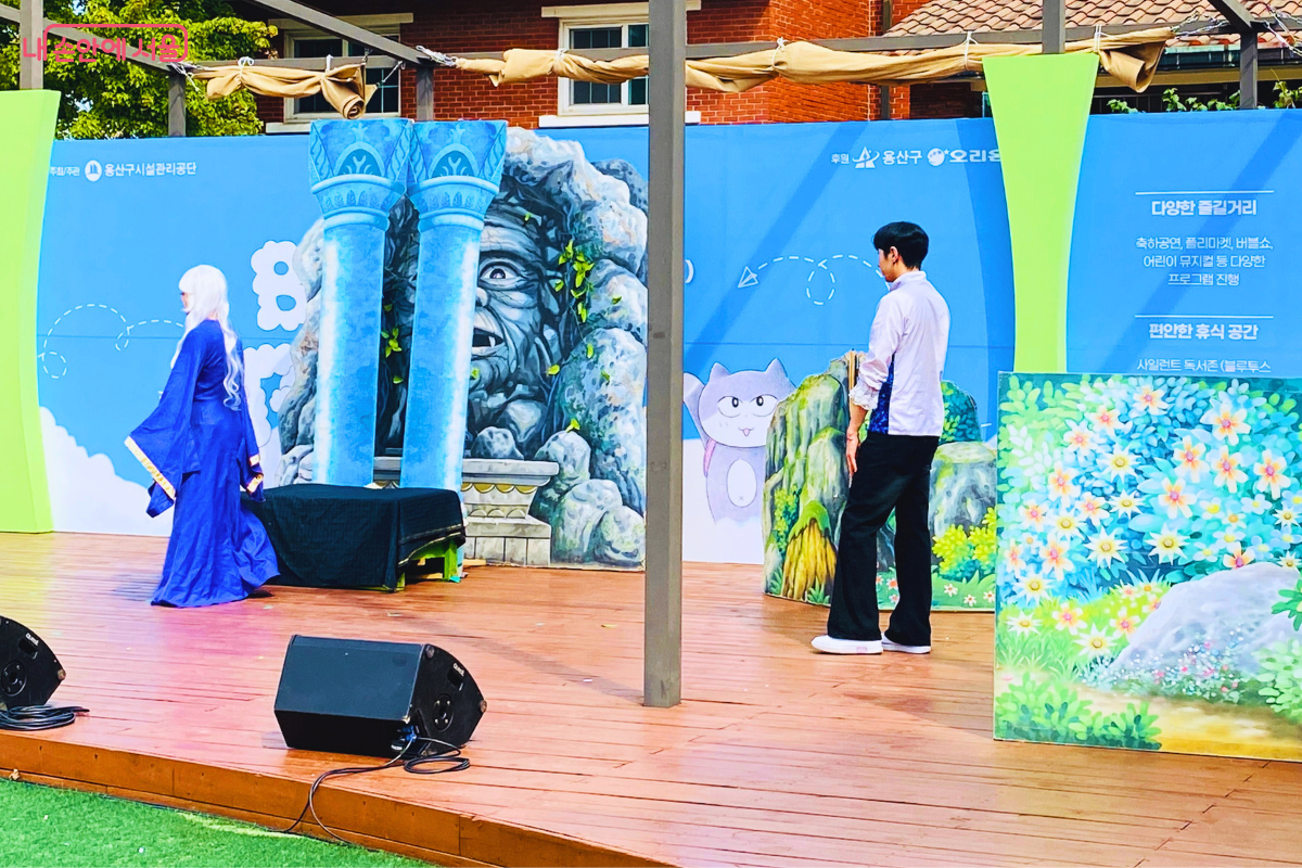야외에 설치된 나무 무대 위에서 어린이 뮤지컬이 진행 중이다. 파란색 드레스를 입은 배우와 남자 배우가 화려한 배경 앞에서 연기를 펼치고 있다. ©양정화