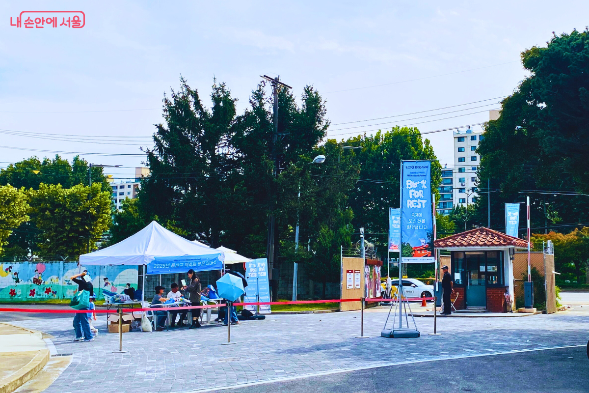 2025 용산구 책축제 '북포레스트'가 열리는 용산공원 장교숙소 5단지 입구 전경. 방문객 안내를 위한 천막과 'Book for Rest'라고 적힌 파란색 배너들이 세워져 있어 축제의 시작을 알리고 있다. ©양정화