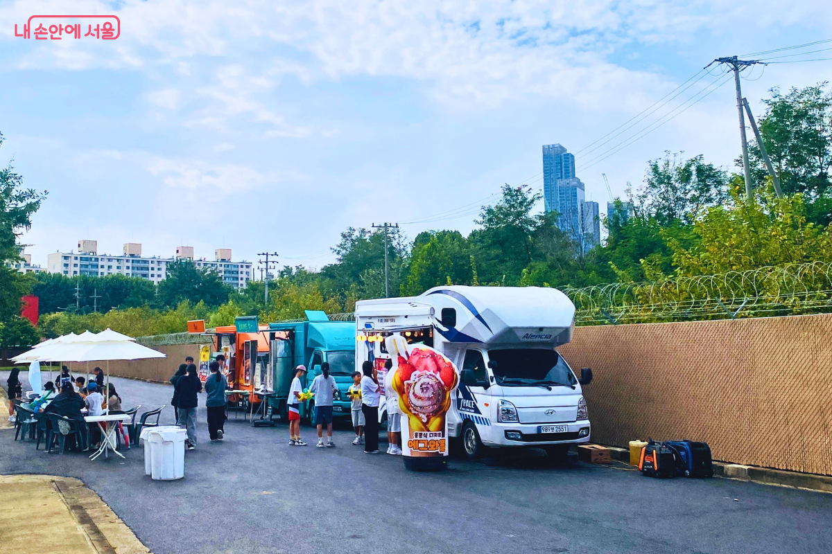 행사장 한쪽에 마련된 푸드트럭 존. 여러 대의 푸드트럭과 캠핑카가 나란히 서 있고, 시민들이 음식을 주문하거나 파라솔이 있는 테이블에서 음식을 먹고 있다. ©양정화
