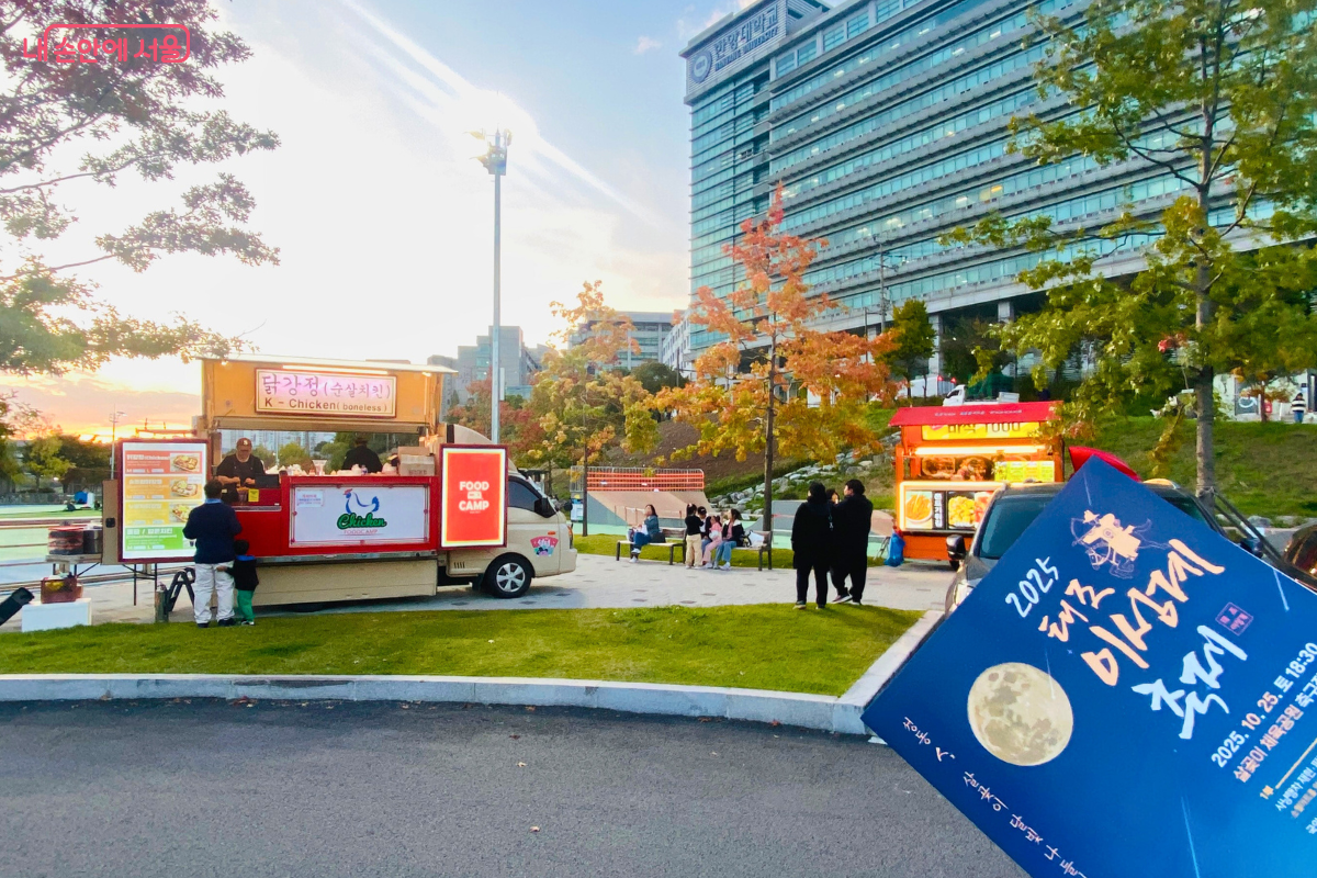 축제가 시작되기 전, 해 질 녘 살곶이 체육공원 한쪽에 마련된 푸드트럭 존의 모습. 닭강정 푸드트럭 등이 영업을 준비하고 있으며, 시민들이 벤치에 앉아 휴식을 취하고 있다. 사진 오른쪽 하단에는 축제 안내 책자가 보인다.