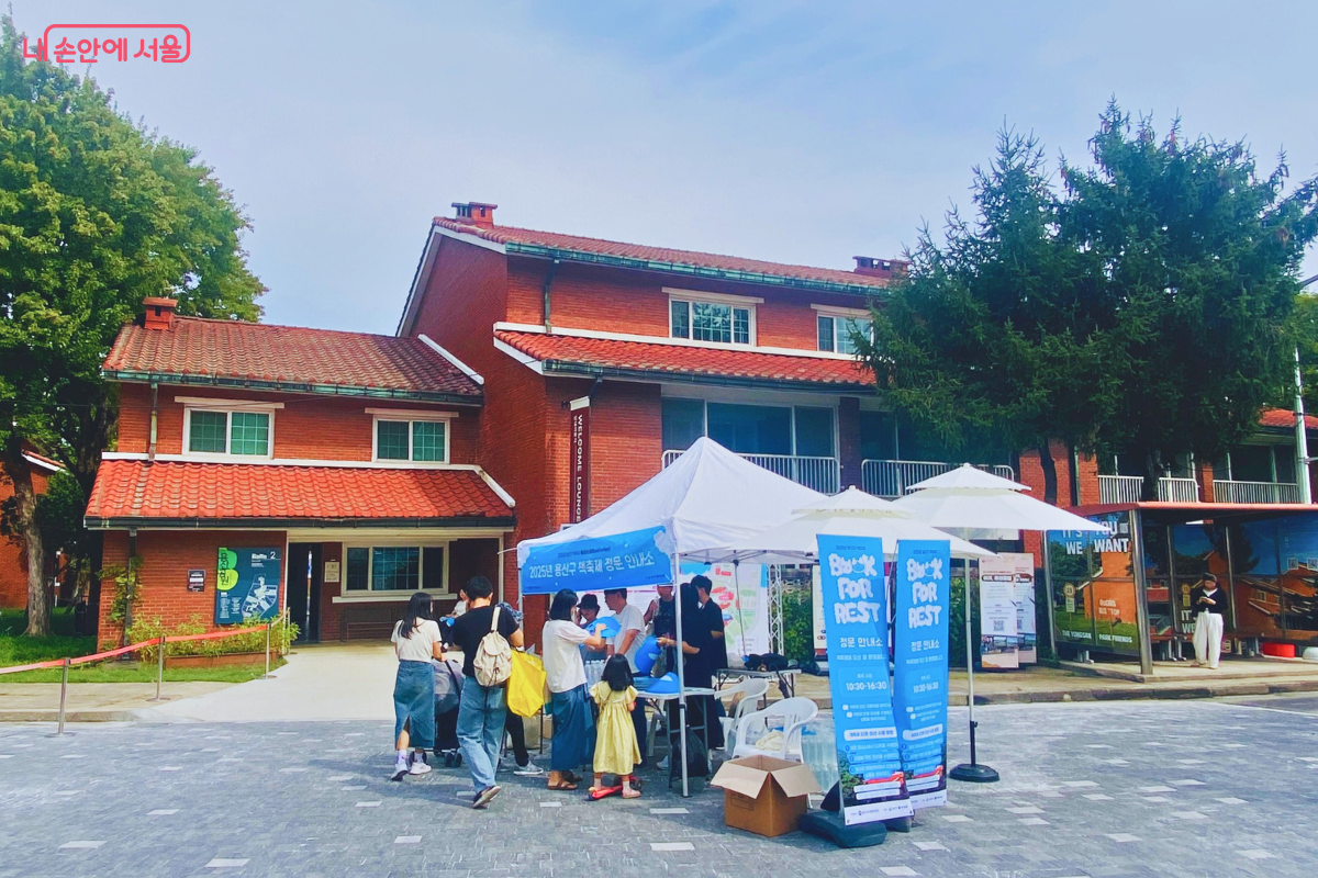'Book for Rest' 배너가 세워진 축제 정문 안내소의 모습. 붉은 벽돌 건물을 배경으로 스태프들이 천막 아래에서 방문객들에게 축제 안내를 하고 있다. ©양정화