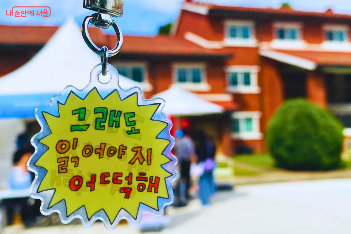 "그래도 읽어야지 어떡해"라는 재치 있는 문구가 적힌 노란색 키링을 클로즈업한 사진. 배경으로 축제가 열리는 붉은 벽돌 건물과 부스가 흐릿하게 보인다. ©양정화