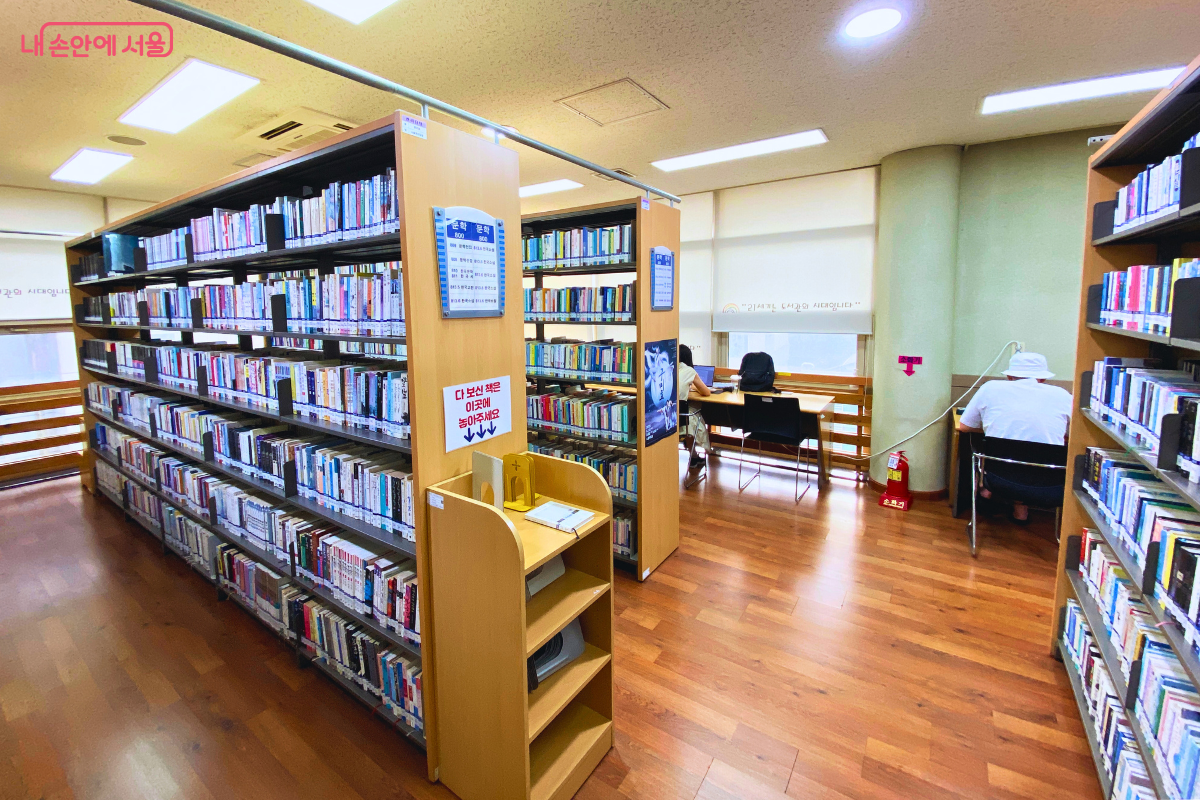 3층 문헌정보실 내부의 모습이다. 책이 가득 꽂힌 높은 서가들 사이로, 밝은 창가에 긴 책상이 '창가 열람석'으로 마련되어 있다. 두 명의 이용객이 창밖을 바라보는 방향으로 자리에 앉아 독서나 공부를 하고 있다. ©양정화