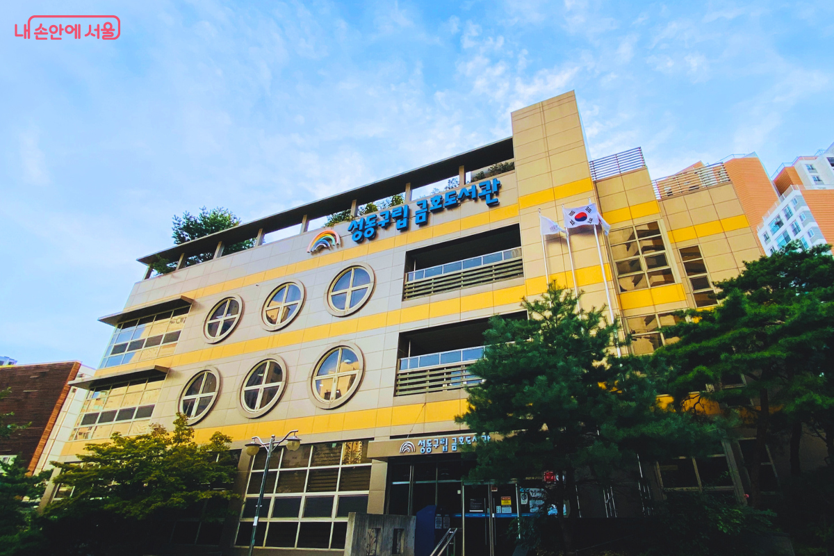 맑고 푸른 하늘을 배경으로 성동구립 금호도서관 건물을 아래에서 올려다본 전경이다. 건물은 5층 규모로, 노란색 수평 띠와 회색 벽이 교차하며 독특한 원형 창문이 특징이다. 상단에는 '성동구립 금호도서관' 간판이, 오른쪽에는 태극기가 게양되어 있다. ©양정화