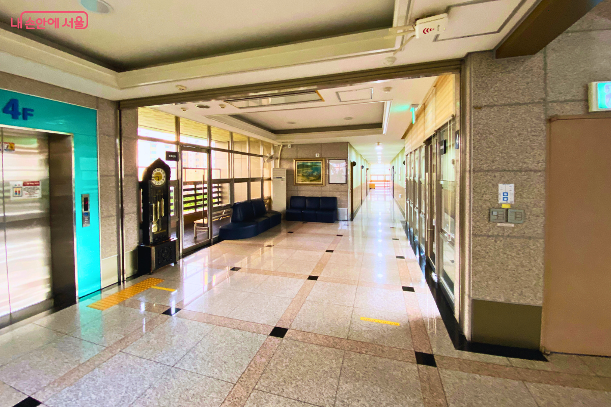 금호도서관 4층의 넓고 밝은 엘리베이터 홀 전경이다. 왼쪽에는 청록색 엘리베이터 문이 있고, 그 옆 휴식 공간에 고풍스러운 괘종시계와 소파가 놓여 있다. 광택이 나는 대리석 바닥 복도가 안쪽으로 길게 이어진다. ©양정화