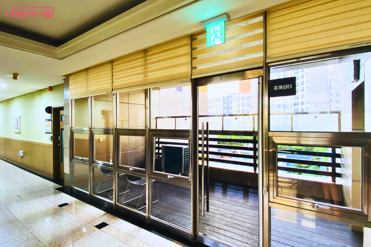 도서관 복도에 마련된 테라스형 '휴게 쉼터'의 입구이다. 통유리창과 문으로 복도와 분리되어 있으며, 안쪽으로 나무 바닥이 깔린 발코니 공간과 바깥 풍경이 보인다. 문 위에는 녹색 비상구 표시등이 켜져 있다. ©양정화