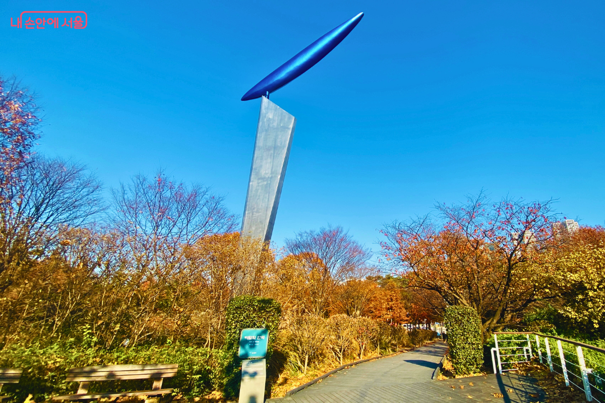 구름 한 점 없는 파란 가을 하늘을 배경으로, 은색의 길고 날렵한 조형물이 솟아 있는 '바람의 언덕' 풍경이다. 주변에는 붉은색과 갈색으로 물든 가을 나무들이 우거져 있고, 잘 조성된 나무 데크 산책로가 오른쪽으로 이어져 있다. ©양정화