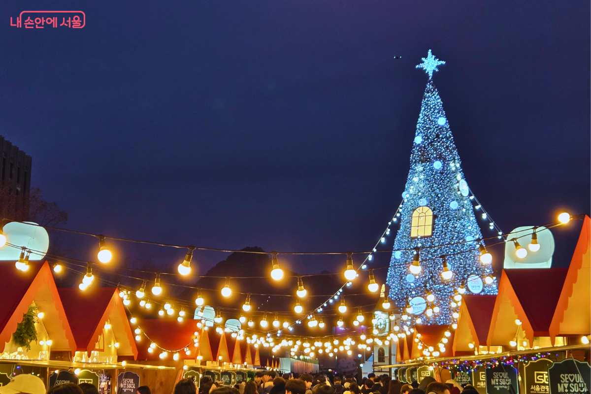 어둠이 내린 광화문광장 마켓의 전경이다. 오른쪽에는 거대한 푸른색 대형 트리가 빛나고, 머리 위로는 따뜻한 색감의 알전구가 줄지어 켜져 있다. 붉은 지붕의 마켓 부스 사이로 수많은 시민이 이동하고 있다.