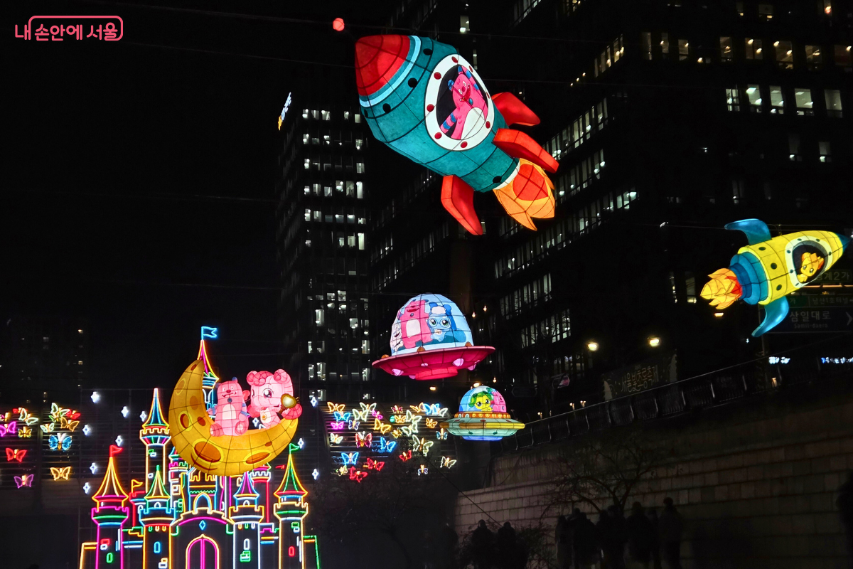 청계천 4구역 상공에 매달린, 서울의 상징 캐릭터 해치와 티니핑 캐릭터를 활용한 우주여행 테마의 유등들. 초록색 로켓에는 해치 캐릭터가, 분홍색 달 모양의 유등에는 티니핑 캐릭터들이 앉아 있으며, 우주선 모양의 다른 등불들도 함께 날고 있다. 아래에는 화려한 색상의 성 모양 조형물과 나비 모양의 조형물들이 배치되어 어린이들에게 꿈과 상상력을 선사하는 듯하다.