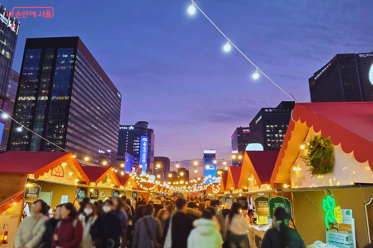 해 질 녘 보랏빛 하늘 아래 광화문 마켓이 수많은 인파로 북적이고 있다. 머리 위로 줄지어 달린 알전구가 거리를 밝히고, 양옆으로 늘어선 붉은 지붕의 마켓 부스 사이를 시민들이 가득 메우며 걷고 있다.