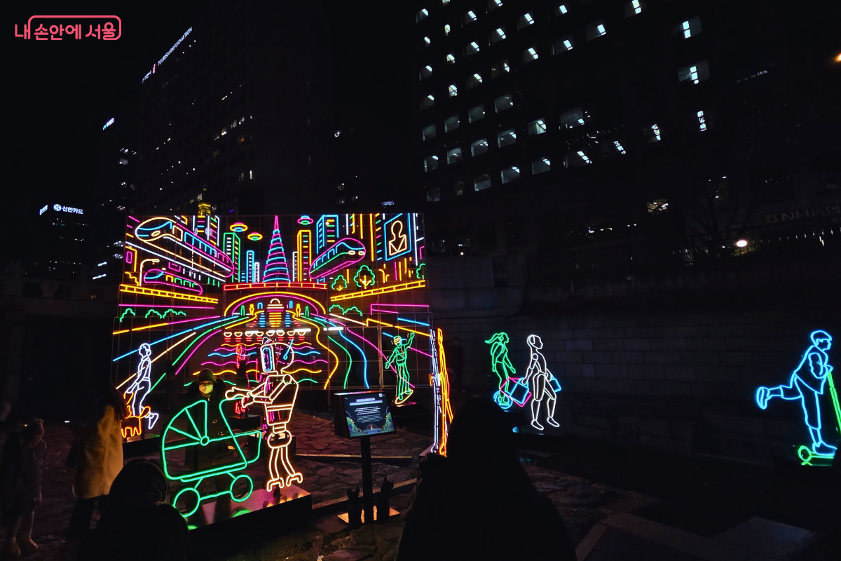 청계천 1구역에 설치된, 미래 도시의 모습을 네온사인 스타일로 표현한 조형물. 밝은 네온 색상(핑크, 청록, 노랑 등)으로 이루어진 스케치 스타일의 등불 조형물이 미래의 청계천과 서울의 스카이라인을 묘사하고 있다. 화면에는 높은 건물, 다리, 지하철 등이 보이며, 전경에는 로봇이나 유모차를 끄는 사람 등 미래의 일상생활 모습이 표현되어 있다. 이는 '청계의 빛, 청계천의 미래'를 주제로 첨단 기술과 조화로운 삶을 상징한다.