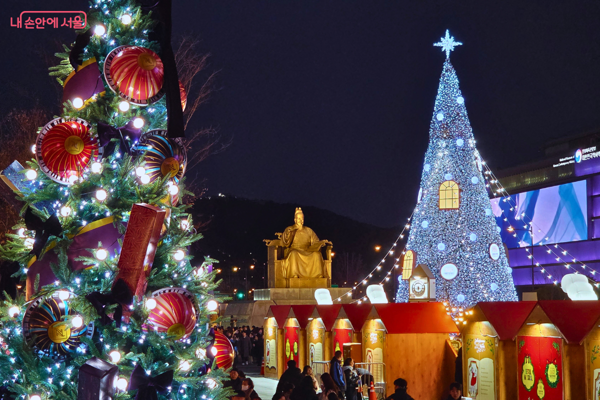 광화문광장의 세종대왕 동상을 중심으로 양옆에 트리가 배치된 모습이다. 오른쪽에는 푸른빛의 메인 대형 트리가, 왼쪽에는 크리스마스 분위기로 장식된 트리가 있어 전통과 현대의 조화를 보여준다.