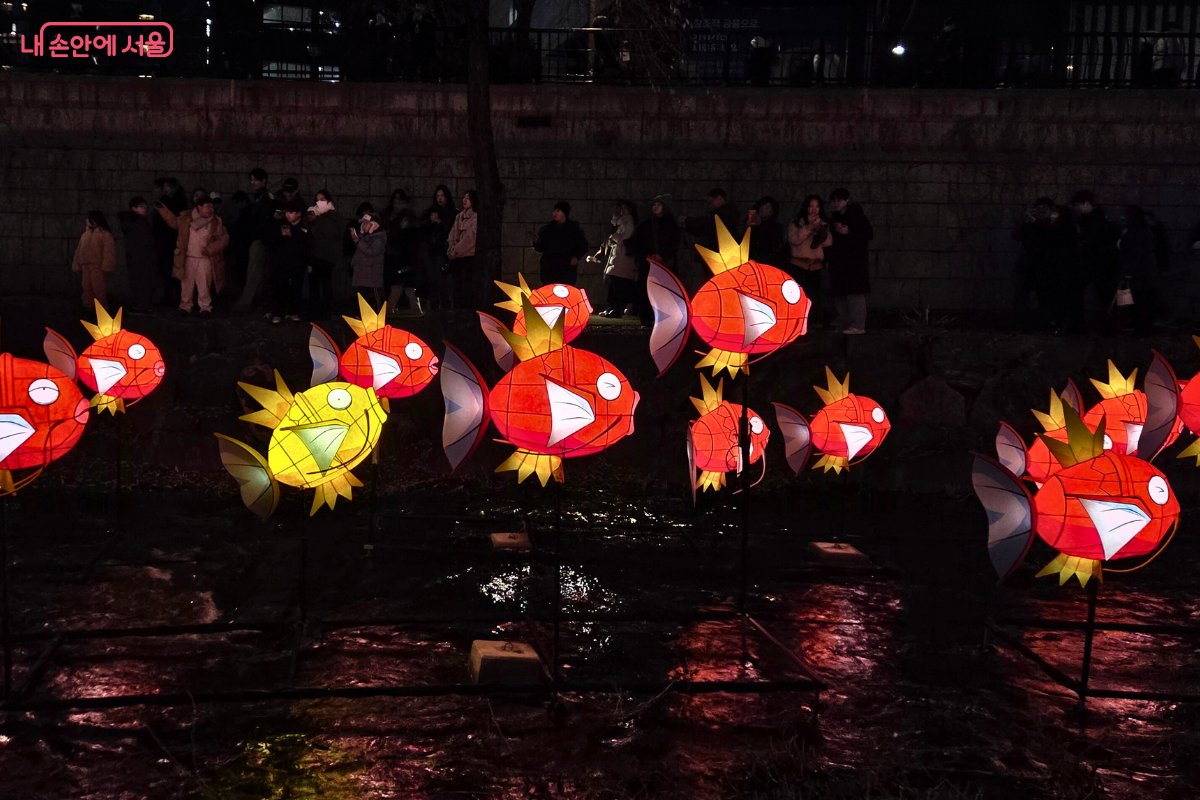 청계천 3구역에 설치된, 포켓몬 캐릭터 '잉어킹(Magikarp)' 물고기 모양의 유등 조형물. 주황색과 노란색이 섞인 몸체와 흰색 입을 가진 단순화된 형태의 물고기 등불들이 청계천 물 위를 따라 무리 지어 떠다니는 모습이다. 조형물 주변의 어두운 배경과 대비되어 밝게 빛나며, 뒤쪽 난간 위에는 축제를 관람하는 시민들의 모습이 보인다.