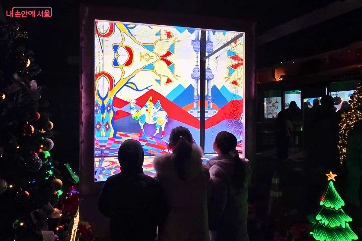 광교 아래 공간에서 대형 스크린에 상영되는 미디어 아트를 관람하는 세 명의 어린이 뒷모습. 화면 속에는 강렬한 색채의 스테인드글라스 스타일로 추상적인 문양과 함께 낙타 등의 동물, 그리고 이국적인 건축물이 표현되어 있다. 화면 양옆과 아래쪽으로는 크리스마스 트리와 별 모양 조형물이 놓여 있어 축제 분위기를 더한다.