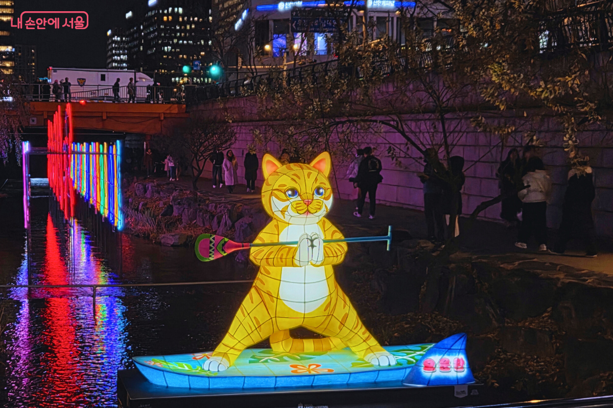 청계천 2구역에 전시된 MZ세대의 트렌드를 반영한 듯한 유등 조형물. 노란색 바탕의 커다란 고양이 캐릭터가 서핑 보드 위에 서서 패들을 들고 요가 자세를 취하는 듯한 모습으로 제작되었다. 캐릭터 아래의 서핑 보드는 푸른색과 흰색 물결 무늬로 꾸며져 있으며, 뒤쪽에는 청계천 물에 비치는 무지개색의 세로 조명 기둥들이 밝게 빛나고 있다.