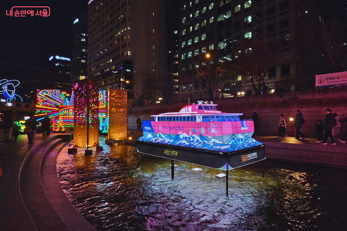 청계천 3구역에 설치된 '한강 리버버스(Hangang Bus)'를 형상화한 대형 유등 조형물. 분홍색과 파란색으로 칠해진 배 모양의 유등이 물 위에 떠 있으며, 하단에는 'Hangang Bus'라는 영문 문구가 적혀 있다. 배의 측면에는 역동적인 물결 무늬가 디자인되어 있다. 주변에는 노란색과 주황색으로 빛나는 탑 모양의 다른 조형물들이 함께 배치되어 있다.