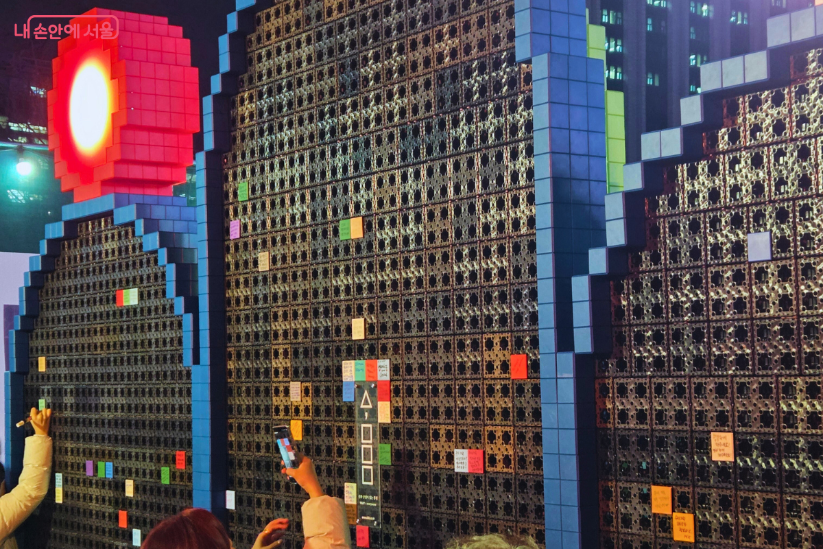 일월오봉도를 형상화한 조형물에 작은 구멍이 뚫려 있고, 시민들이 알록달록한 메모지 블록에 소원을 적어 촘촘하게 붙이고 있다. 왼쪽 위에는 붉은색 픽셀 원형 해 모양 조명이 빛나고 있으며, 시민들이 참여하여 완성하는 빛 조형물의 모습이다.