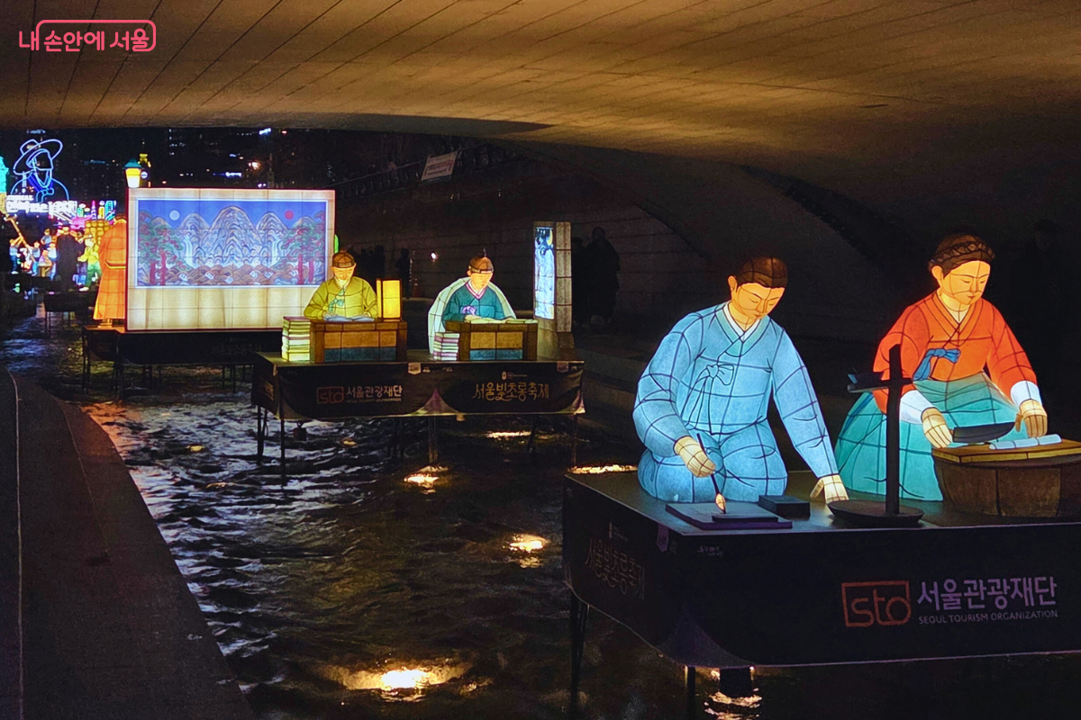 청계천 다리 아래 어두운 공간에 설치된 조선시대 선비들의 일상생활을 재현한 전통 등불(유등)의 모습. 책상에 앉아 독서하거나 글씨를 쓰는 두 명의 선비와, 돗자리 위에 앉아 바느질 또는 다듬이질을 하는 듯한 부녀자 두 명의 유등이 물 위에 떠 있다. 유등들은 은은한 노란빛과 푸른빛을 내며, 뒤쪽의 대형 배경 등에는 산수화가 그려져 있어 고즈넉한 옛 분위기를 연출한다.