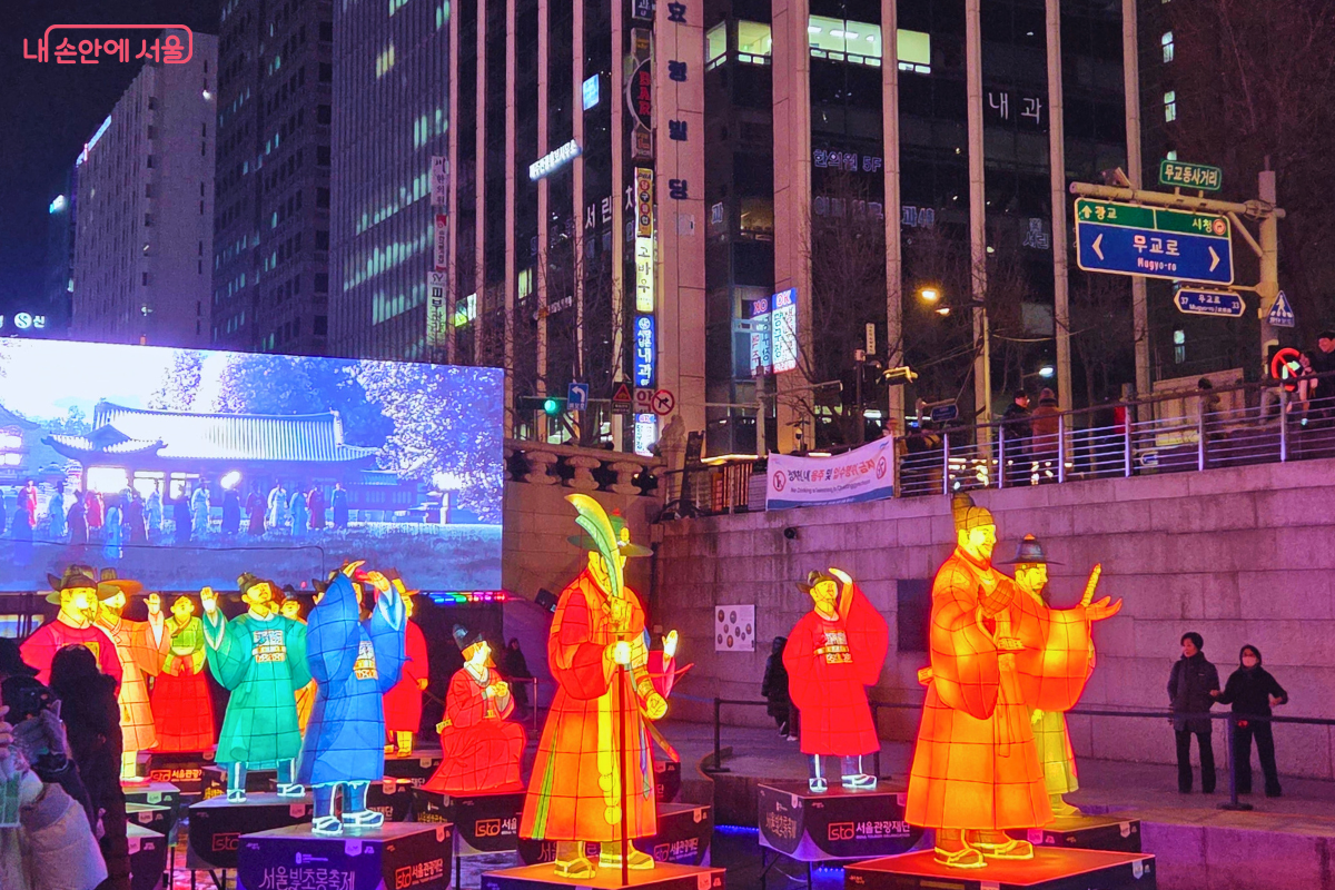 청계천 1구역에서 밤을 밝히는 전통 등불 조형물의 모습. 조선시대 왕과 신하 또는 무관 복식을 한 인물 형상의 대형 유등들이 청계천 변에 설치된 단상 위에 세워져 있다. 유등들은 따뜻한 노란색 빛을 중심으로 붉은색, 청록색, 보라색 등으로 다채롭게 빛나고 있으며, 뒤편 대형 스크린에는 궁궐의 야경이 영상으로 상영되고 있다. 