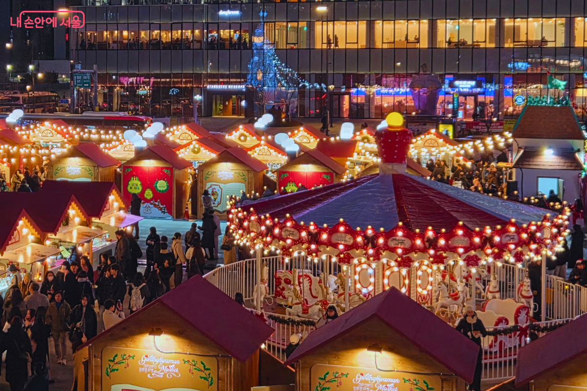 위에서 내려다본 마켓의 풍경이다. 오른쪽 앞에는 회전목마의 지붕이 크게 보이고, 그 주변으로 붉은색 세모 지붕을 한 먹거리 부스들이 옹기종기 모여 있다. 부스마다 따뜻한 조명이 켜져 있고, 시민들이 줄을 서거나 음식을 즐기고 있다.