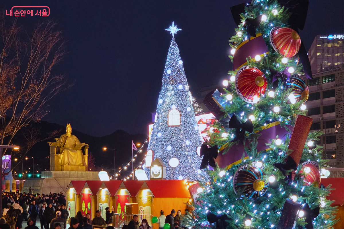 화면 앞쪽에는 화려한 오너먼트와 리본으로 장식된 크리스마스 트리가 크게 보이고, 그 뒤편으로 푸른 조명의 대형 트리와 황금빛 세종대왕 동상이 나란히 자리하고 있다. 밤하늘 아래 다양한 조명이 어우러져 웅장한 분위기를 자아낸다.