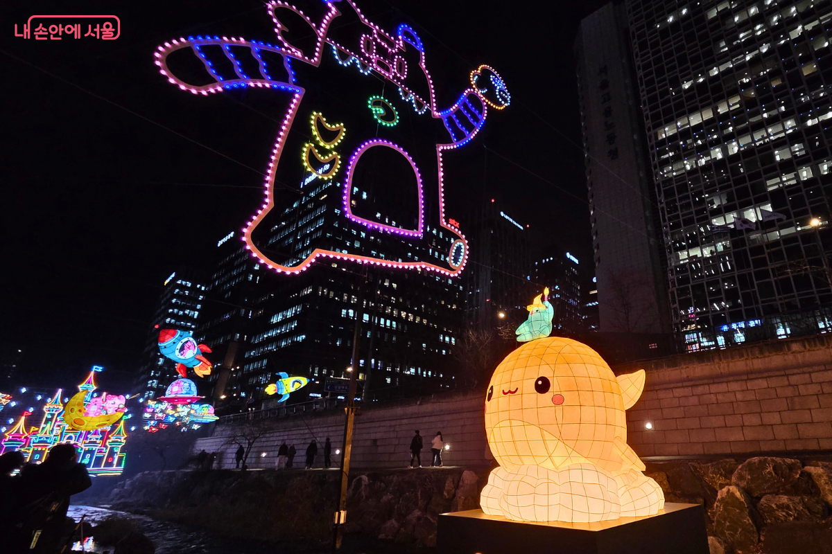 청계천 4구역에 설치된 '서울 판타지아' 구역의 다양한 캐릭터 유등 조형물. 정면에는 노란색과 주황색의 귀여운 아기 고래 유등이 밝게 빛나고 있다. 그 위쪽으로는 서울의 상징 캐릭터인 해치와 인기 애니메이션 캐릭터 티니핑이 우주선 및 로켓을 타고 하늘을 유영하는 모습의 대형 등불들이 매달려 있다. 왼쪽 아래에는 화려한 색상의 성 모양 조형물이 배치되어 환상적인 분위기를 연출한다.