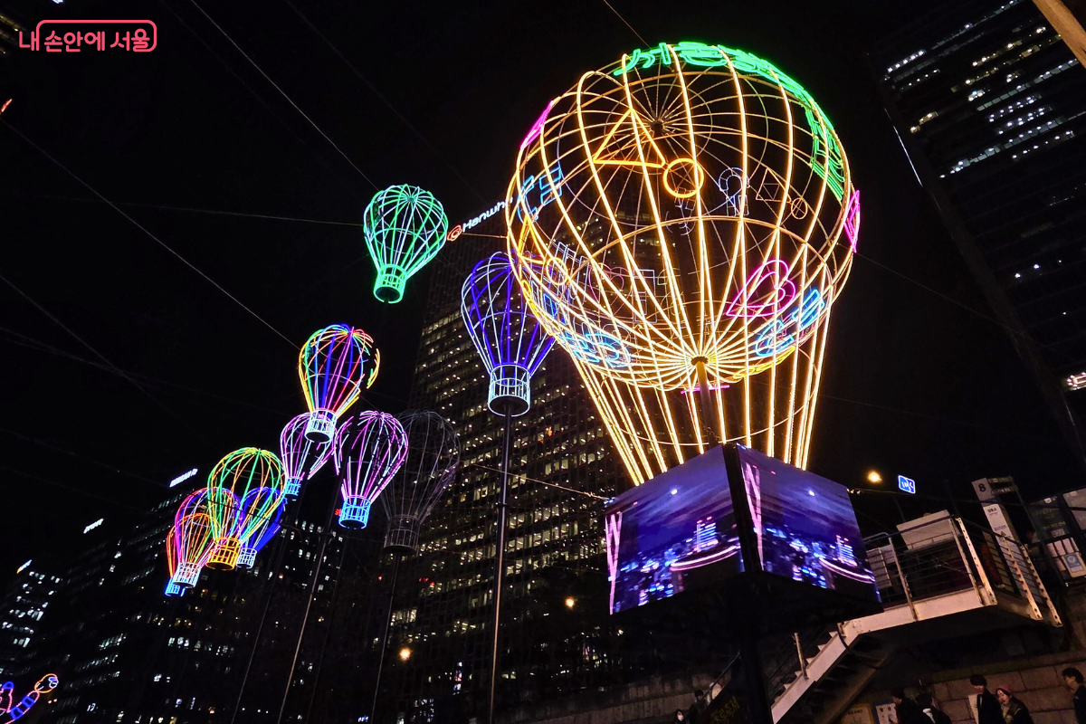 청계천 4구역 상공에 매달린 여러 개의 열기구 모양 LED 조형물. 가장 중앙의 대형 열기구는 노란색과 주황색 계열의 조명으로 빛나고, 주변의 작은 열기구들은 청록색, 보라색, 무지개색 등 다채로운 색상으로 빛난다. 조형물 아래에는 영상을 상영하는 듯한 입방체 형태의 스크린이 설치되어 있으며, 뒤쪽에는 고층 빌딩들의 야경이 배경을 이룬다.