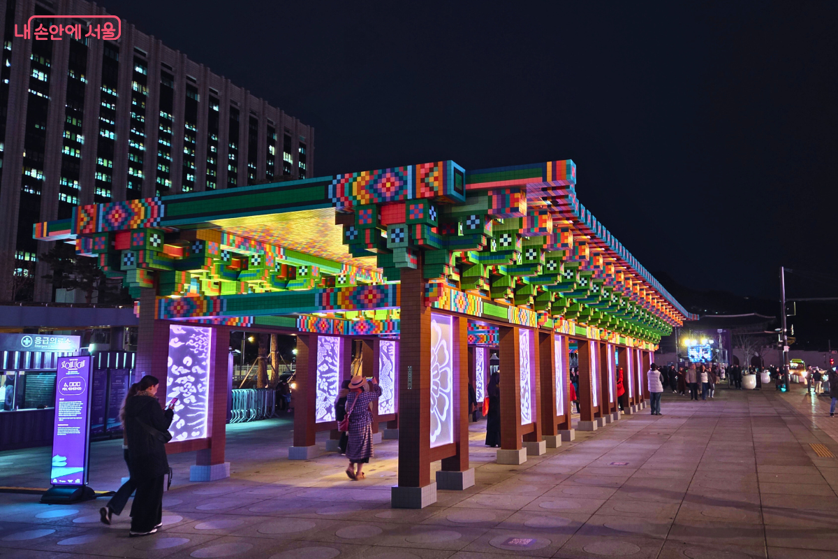 전통 단청 무늬를 알록달록한 픽셀 아트로 재해석한 긴 회랑 형태의 빛 조형물이다. 화려한 색감의 블록으로 만들어진 기둥과 지붕이 밤거리를 밝히고 있으며, 시민들이 그 아래를 거닐거나 사진을 찍고 있다.