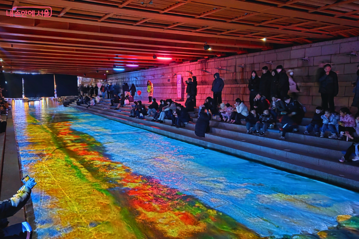  광교 아래 청계천 바닥에 투사된 몰입형 미디어 아트 '청계 소울 오션'의 모습. 청계천 물 위에 강렬한 노란색과 주황색, 그리고 시원한 푸른색과 청록색이 혼합되어 마치 바다나 우주 공간처럼 역동적으로 움직이는 빛을 연출하고 있다. 주변에는 붉은 조명이 비추는 교량 하부 구조물과 계단에 앉아 이 광경을 관람하는 많은 시민들의 모습이 보인다.