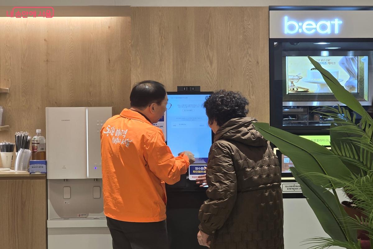 키오스크 앞에서 봉사자의 설명을 듣고 있는 어르신 ©강사랑