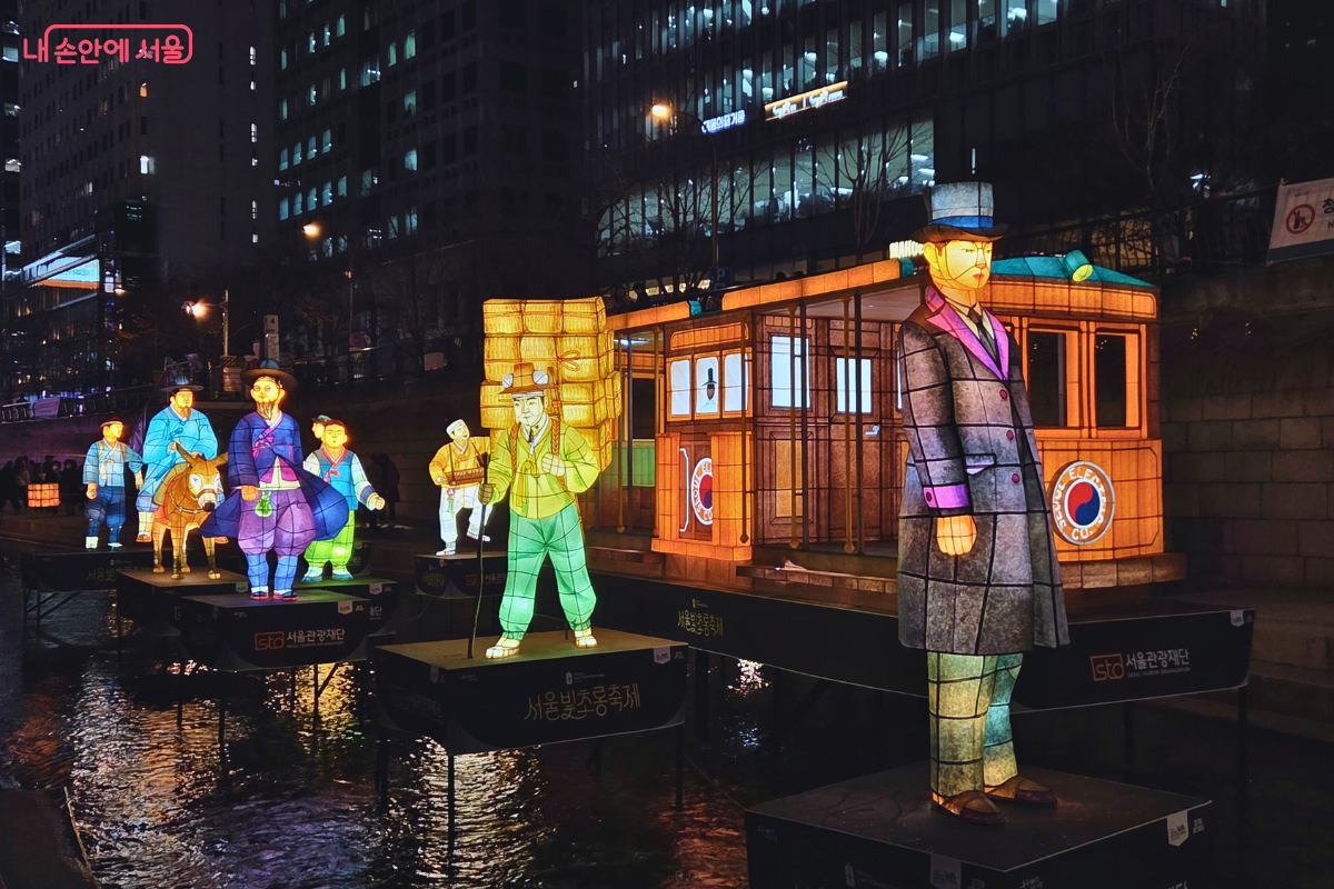 청계천 1구역에 전시된 근대 개화기 서울의 모습을 담은 전통 등불 조형물. 전차(트램) 형태의 대형 등불을 중심으로, 서양식 정장을 입은 신사와 한복을 입은 인물, 그리고 나귀를 탄 인물 등 다양한 복식의 사람들이 함께 있는 모습을 표현하고 있다. 이는 근대 문물이 유입되던 시기의 서울 거리를 재현한 것으로, '움직이는 빛'이라는 주제로 다양한 교통수단과 복식을 보여준다. 유등들은 다채로운 색상으로 빛나며 과거와 현재가 공존하는 모습을 보여준다.